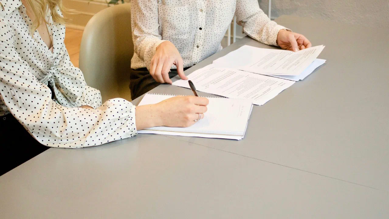 A person signing a printed document while another points at the page
